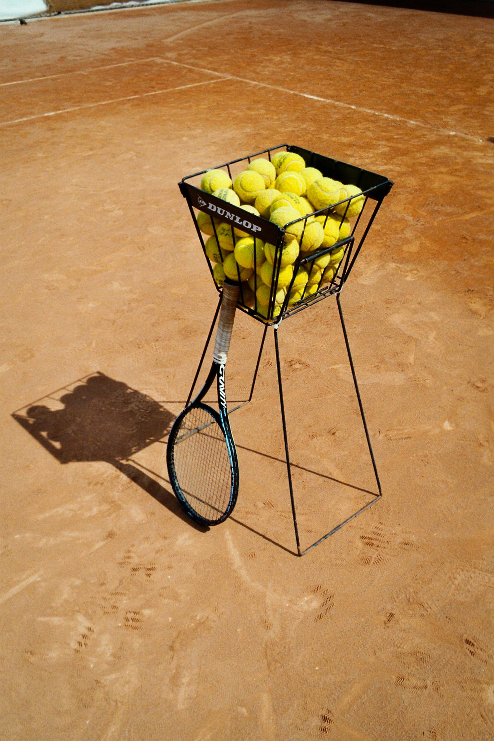 pexels-photo-24194315-24194315 A basket filled with tennis balls and a racket on a clay tennis court.